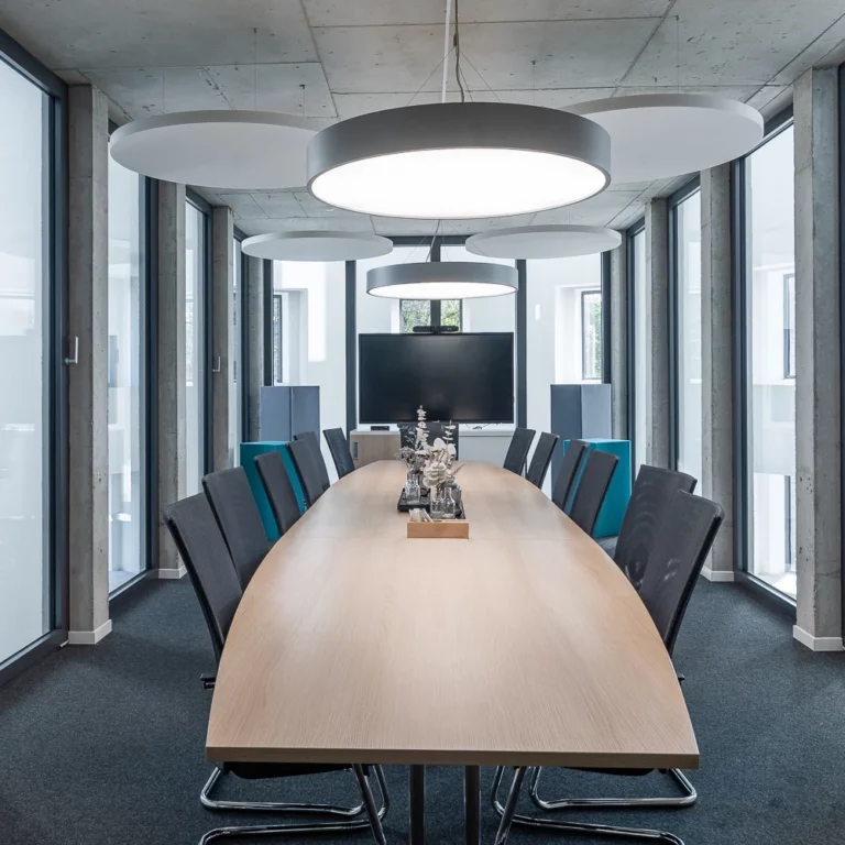 Heller Konferenzraum mit langem Tisch, Bürostühlen und einer Deckenleuchte, im Hintergrund eine Glasfront.
