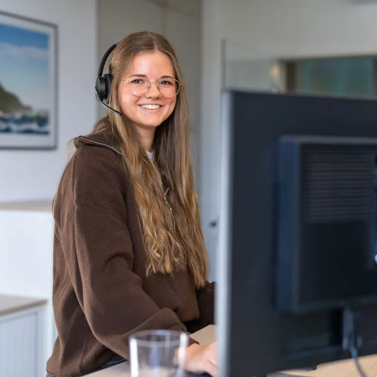 Eine lächelnde junge Frau mit Kopfhörern um den Hals steht in einem hellen Büro vor einer Wand mit Bildern.